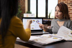 Project manager seated with coworker at desk, pointing to financial graphs on laptop during their business meeting. Asian female advisor describing company analytics to employee in brick wall office.