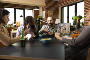 Multiethnic group of friends enjoy card game with drinks and snacks. Male and female individuals chat and laugh while sitting at table with chips and beer during weekend bonding time.
